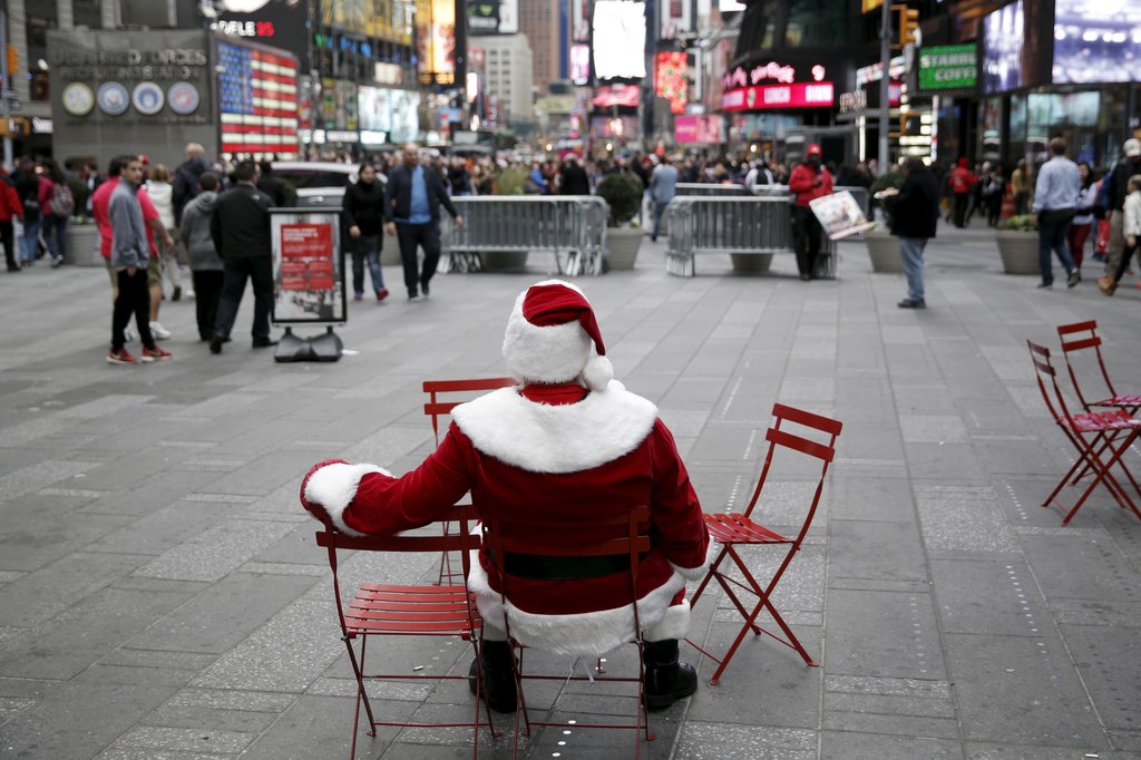 A person dressed as Santa Claus sits alone on a red folding chair in New York City’s Times Square, facing a busy pedestrian area. Surrounded by empty chairs and distant crowds, the Santa figure appears isolated amid the hustle of the city. Bright lights, storefronts, and digital billboards fill the background.